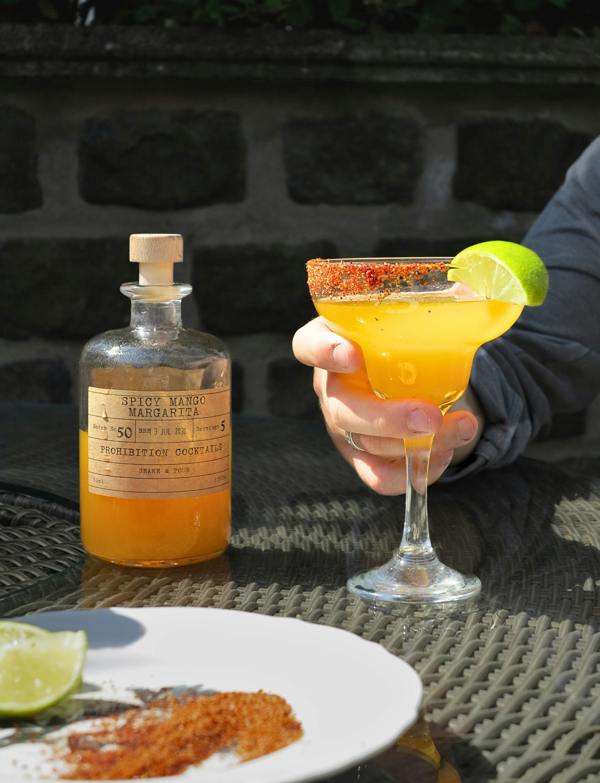 A hand holds a margarita-style cocktail with a lime wheel and chili powder rim in a glass, next to a labeled bottle of orange liquid on a patio table.