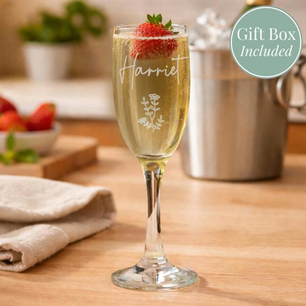 An engraved champagne flute by Abode Home & Gifts filled with prosecco and garnished with a strawberry, standing on a wooden countertop. The glass is custom etched with the name "Harriet" and a wildflower motif. An ice bucket and a green circle reading "Gift Box Included" sit in the background.