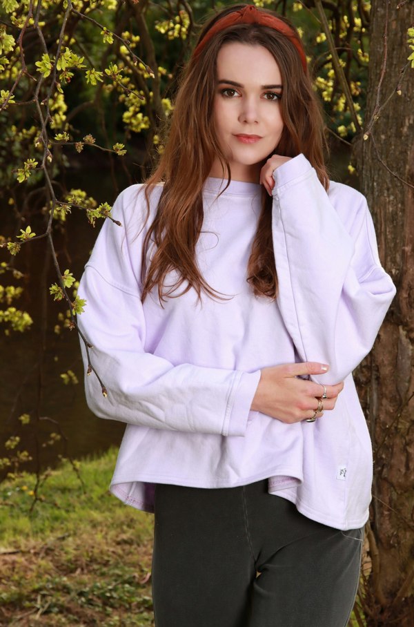 This is a young woman looking at the camera. She is standing in front of a tree which has yellow leaves. She has long hair with a red band. Her left hand is near her chin while her right hand is holding her left elbow. She is wearing a bella oversized cropped sweat shirt in lilac. Here trousers are grey.