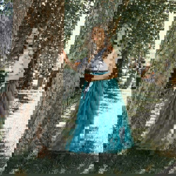 Young girl in a white top and teal skirt standing next to a tree in a park.