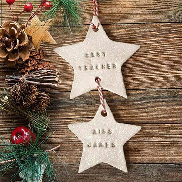 A handcrafted white clay double-star ornament hanging on a wooden background, hand stamped with 'Best Teacher' and 'Miss James'.