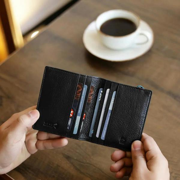 Person holding a black wallet with cards on a wooden table with a cup of coffee in the background.