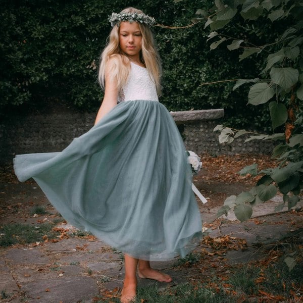 Young girl in a white top and sage green skirt with a flower crown standing outdoors.