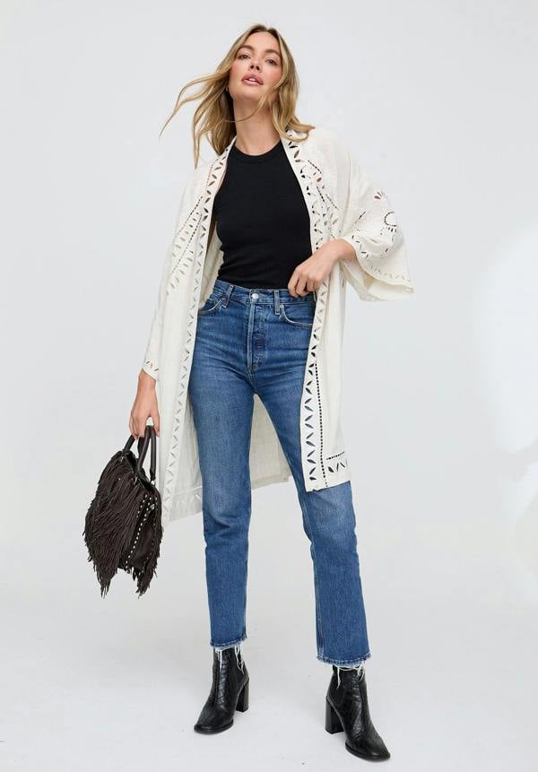 Woman wearing a long white cardigan, black top, blue jeans, and black boots on a white background
