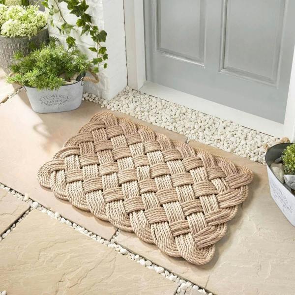 Woven doormat on a stone patio with plants and a door in the background