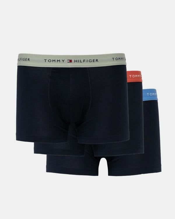 Three pairs of Tommy Hilfiger men's boxer briefs in dark navy blue, stacked together on a light gray background. The front pair has a light gray waistband with the Tommy Hilfiger logo, while the pairs behind it have red, orange, and blue waistbands.