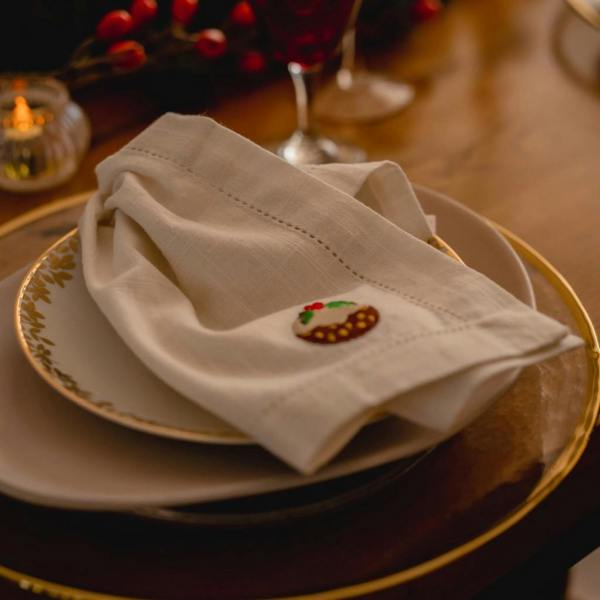 Luxury white cotton napkin with embroidered Christmas pudding motif styled on a gold-rimmed plate for a festive dinner setting.