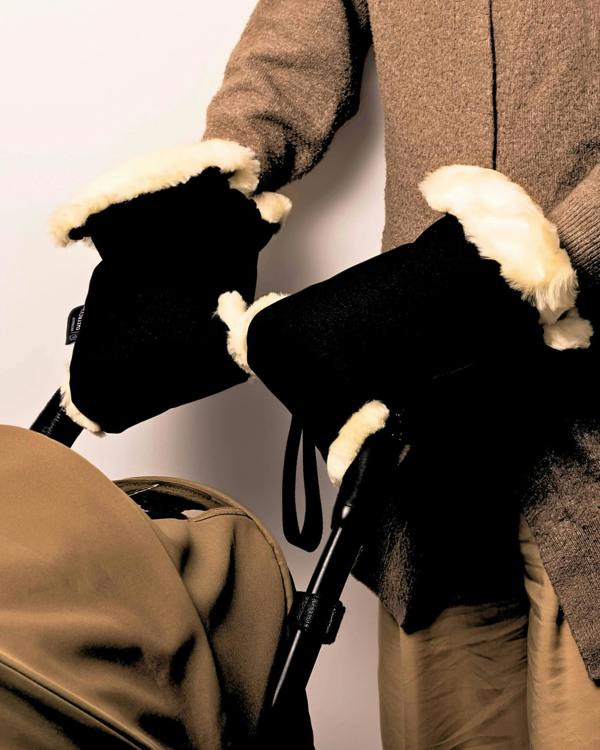 Person wearing black sheepskin gloves with white fur trim holding a brown coat and bag.