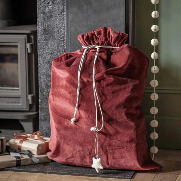 Rich red cotton velvet Christmas gift sack with drawstring closure sitting beside a cosy fireplace hearth surrounded by wrapped presents