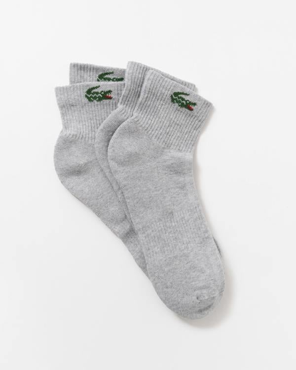 A pair of light gray heather crew socks with the green embroidered Lacoste crocodile logo on the cuff, displayed on a clean white background.