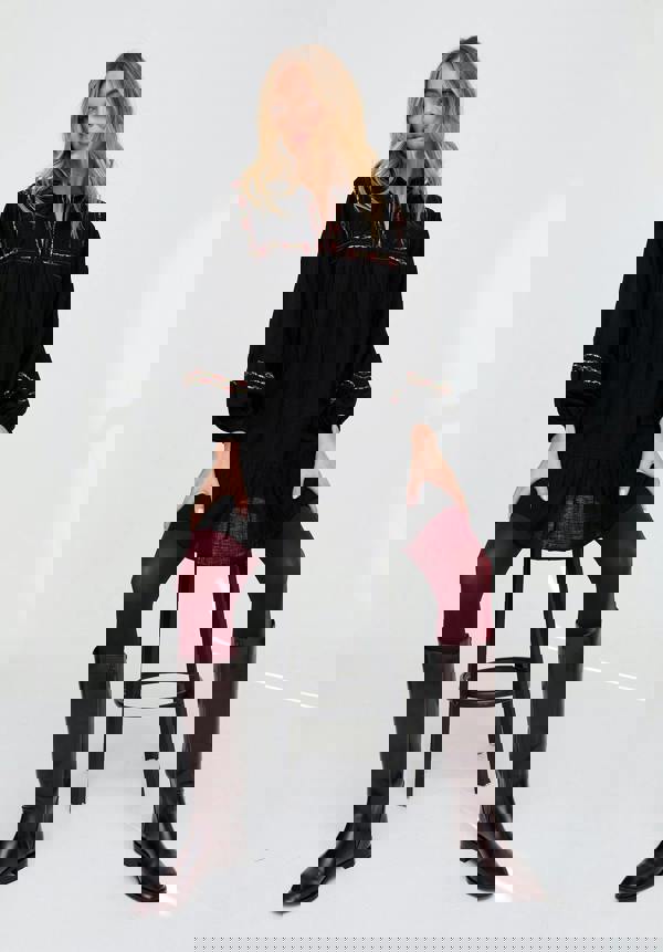 Woman sitting on a stool wearing a black dress with red embroidery, maroon tights, and brown knee-high boots.