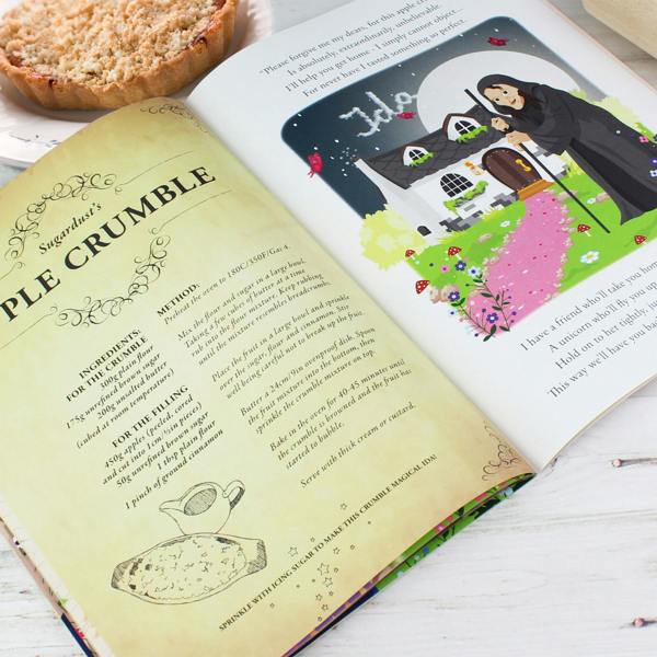 Open view of the Personalised Fairy Baking Adventure storybook showing Sugardust's Apple Crumble recipe beside a story illustration of a witch's cottage with clouds spelling Ida, styled next to a freshly baked crumble tart