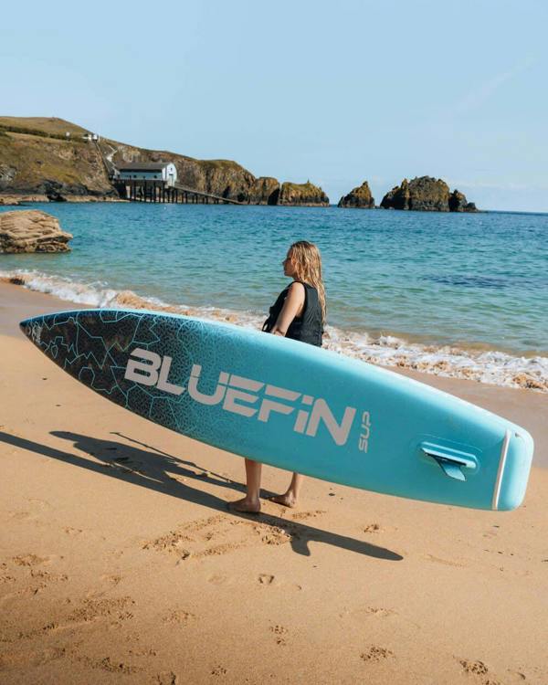Woman carrying a Bluefin SUP on the beach, with clear water and a pier in the background.