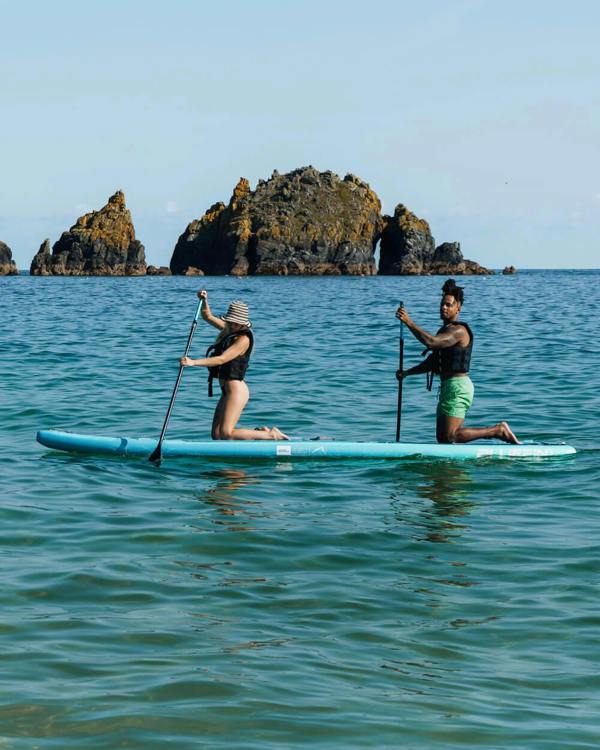 Two people paddle boarding on a calm waterway with rocky formations in the background.