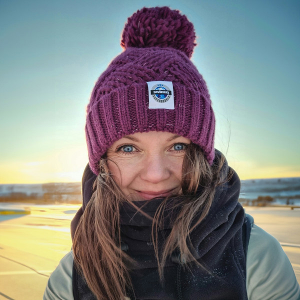 Women in a frozen landscape wearing a hat in deep rhubarb colour with a big bobble. It is knitted in chunky yarn in an attractive cross patch pattern with a deep ribbed headband. The hat contains reflective yarns and is lined in luxury sherpa fleece.