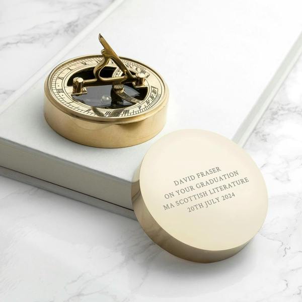 A studio photograph of a polished brass sundial compass resting on a white marble surface with subtle grey veining. The compass lid is positioned in the foreground, clearly engraved with the text "DAVID FRASER ON YOUR GRADUATION MA SCOTTISH LITERATURE 20TH JULY 2024". The compass mechanism is visible in the background, open and reflecting light.