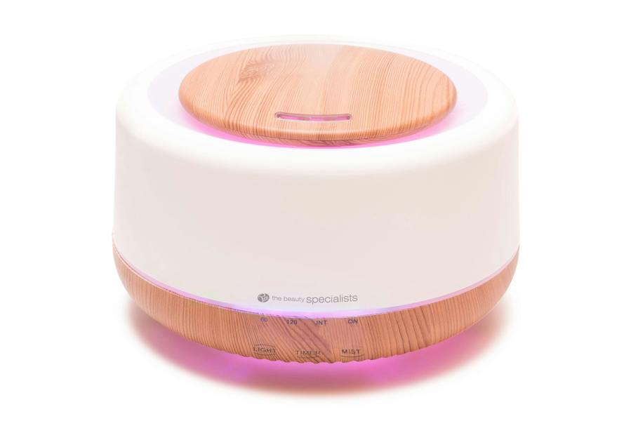 The Rio ALORA Aroma Diffuser, Humidifier & Night-Light features a round design with wood grain top and base, a white middle section, soft pink LED glow, and buttons with labels on the lower front.