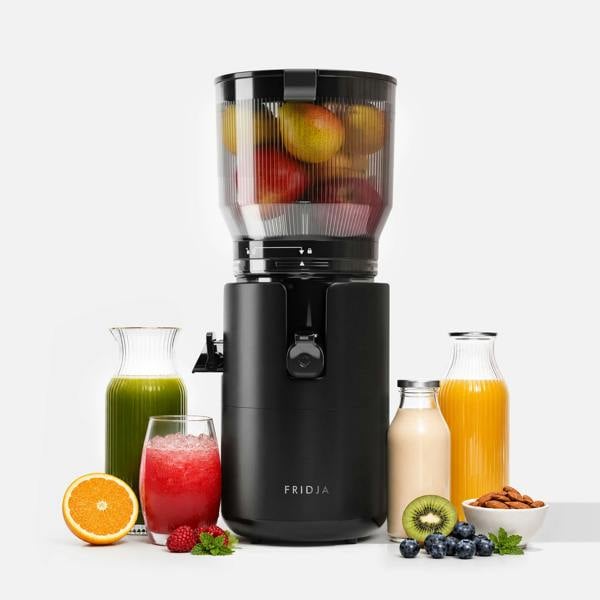 A Fridja f2800 Open Whole Fruit Juicer in black, filled with apples, is surrounded by jars and glasses of green juice, orange juice, a smoothie, fresh oranges, kiwi, blueberries, raspberries, and a bowl of almonds.