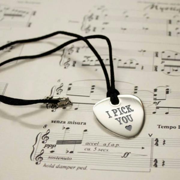 A personalised silver guitar plectrum pendant engraved with 'I PICK YOU' resting on a sheet of classical music notes with a black cord.