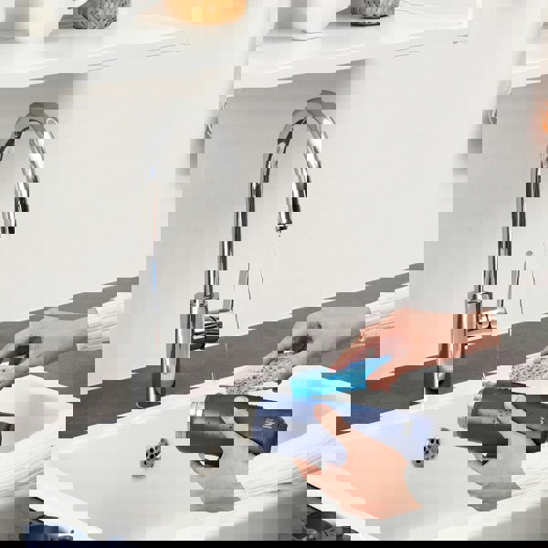 Hands scrubbing a navy bottle with a blue sponge under a running faucet in a kitchen sink context.