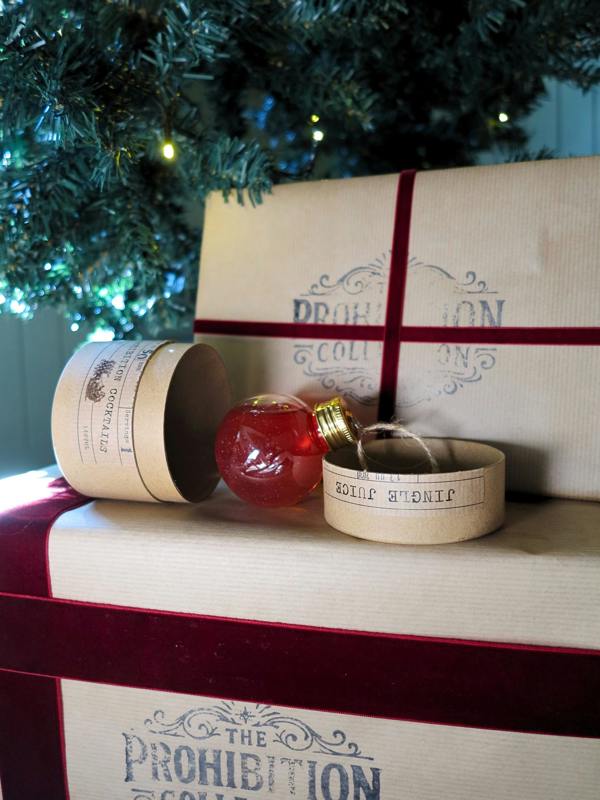 A Christmas-themed gift set from The Prohibition Collection, featuring a red glass ornament, a small paper cylinder, and a larger paper gift box with a dark red ribbon, styled under a decorated Christmas tree.