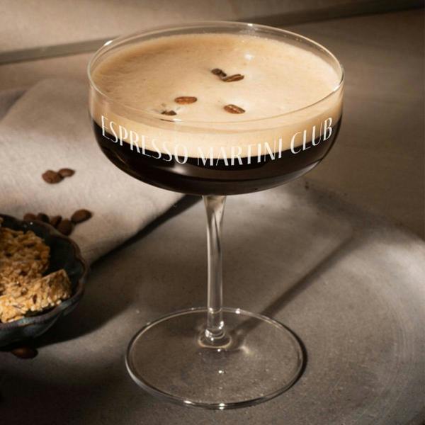 An elegant glass coupe filled with a dark, rich Espresso Martini cocktail, topped with three coffee beans. The glass is custom engraved with the text "ESPRESSO MARTINI CLUB" wrapped around the bowl, resting on a textured grey concrete coaster with a small plate of biscotti nearby.