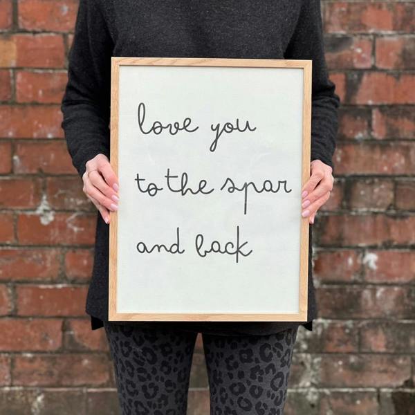 A woman wearing leopard print leggings holding a solid oak framed white cotton typography print displaying the lowercase handwritten-style quote "love you to the spar and back" against a red brick wall.