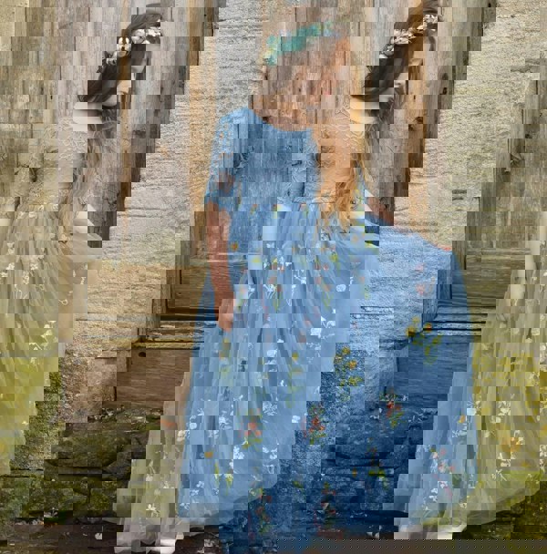 Young girl in a blue floral dress standing against a rustic stone wall.