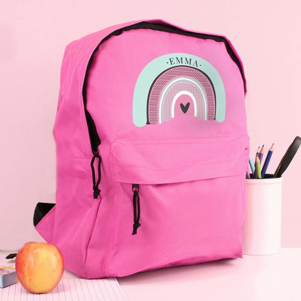 A personalised pink backpack featuring a boho pastel rainbow design and printed with the name EMMA, styled on a desk alongside an apple and a cup filled with pencils.