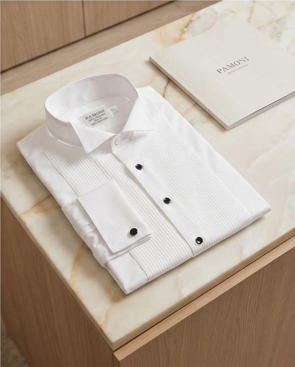 A folded white dress shirt with a subtle vertical texture and black buttons, displayed on a light marble surface next to a branded Pamoni booklet.