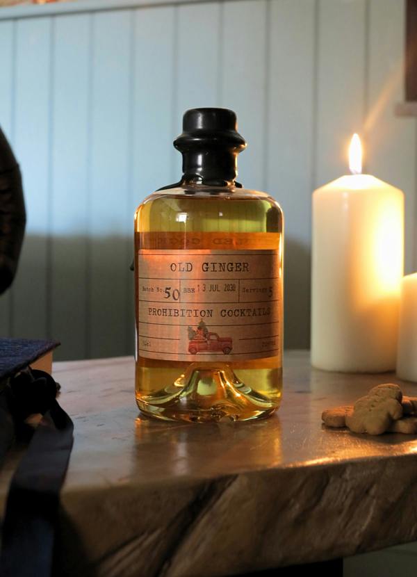 A bottle of Old Ginger Prohibition Cocktails filled with amber liquid, set on a rustic wooden table next to lit white pillar candles and ginger roots.