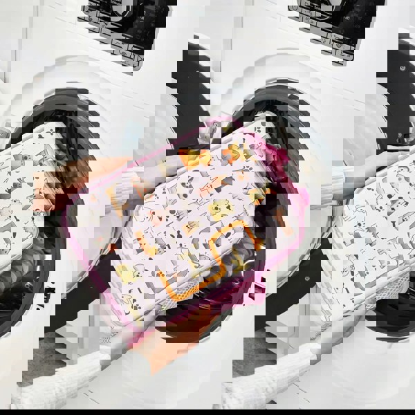 A person places a pink pet-themed zippered pouch with animal illustrations into a front-loading washing machine in a laundry room setting.