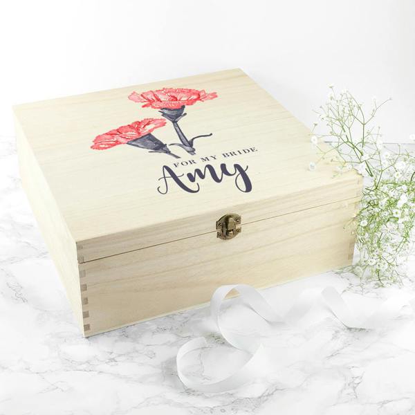 A closed square natural pine box printed with 'For My Bride Amy' and two pink carnations, styled on a white marble surface next to baby's breath flowers and a white ribbon.