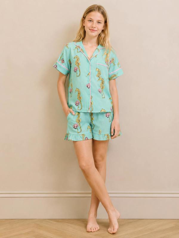 A teenager modelling a two-piece pajama set featuring a light aqua blue base with a whimsical print of cartoon seahorses and small flowers, set against a neutral beige wall and light wood floor.