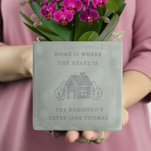 A close-up shot of a square concrete plant pot held by a person in a pink top, featuring an engraved house illustration and personalized text, filled with bright pink orchids.