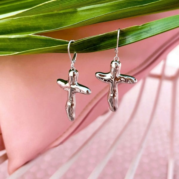 Sterling silver cross earrings hanging off a palm leaf on pink background.