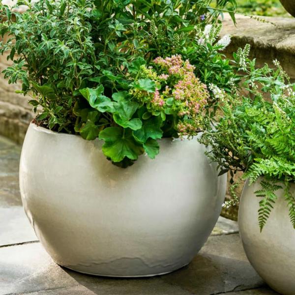 Two round white glazed terracotta planters in varying sizes sitting on a stone patio, filled with lush green garden plants and pink flowers.