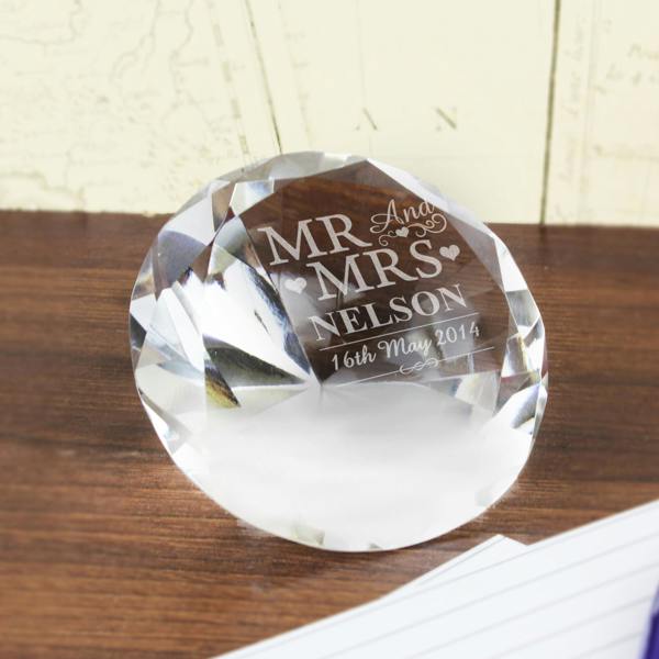 A large, brilliant-cut clear glass diamond paperweight resting on a dark wooden desk. The flat top surface is engraved with "MR AND MRS NELSON" and the date "16th May 2014" in an elegant white font. In the background, vintage-style maps and white lined paper are visible.