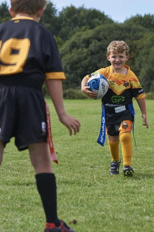 Young player running with ball in blue Optimum tag belt toward a defender in black kit, on grass