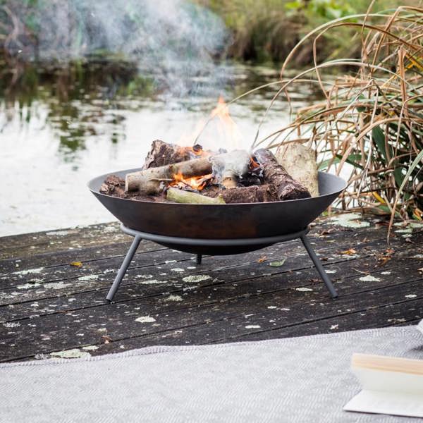 An Idbury Cast Iron Fire Pit with a log fire burning, positioned on a wooden jetty by a pond with a grey throw and book nearby.
