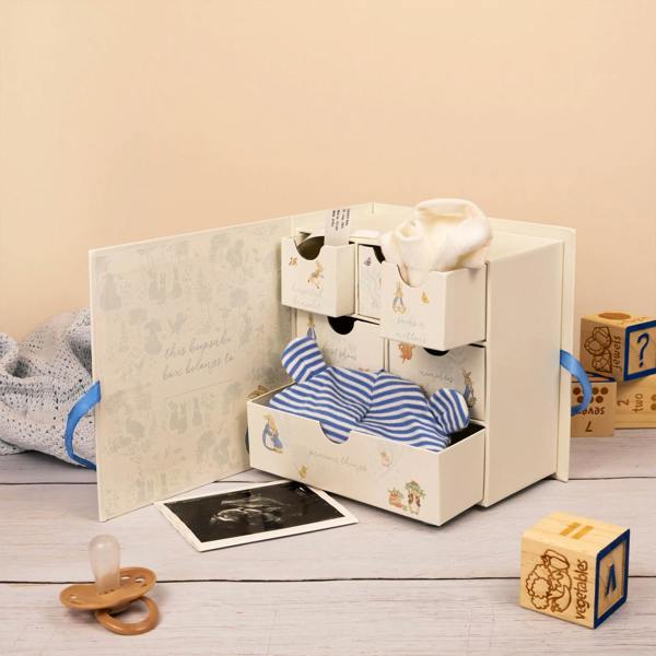 Open Peter Rabbit baby memento chest featuring six drawers labeled for hospital bracelets, socks, and first shoes. The box is decorated with classic Beatrix Potter illustrations, tied with a blue ribbon, and shown holding a baby hat, socks, and an ultrasound photo.
