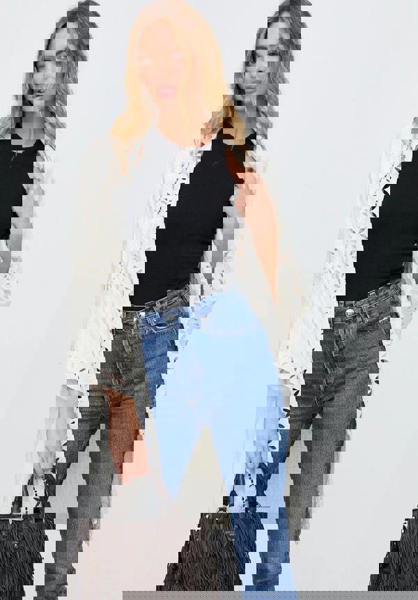 Woman wearing a white lace cardigan, black top, blue jeans, and holding a brown handbag on a light gray background