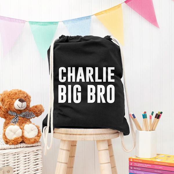 Boy's personalised black gym drawstring bag printed in bold white text with CHARLIE BIG BRO, resting on a wooden stool.