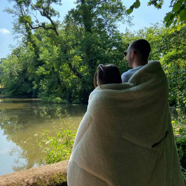 Tonal treetops and vivid spruce Insulated Outdoor Blanko been worn by a young couple enjoying the countryside overlooking a lake with the outdoor blanket, wrapped around them keeping them warm and cosy.