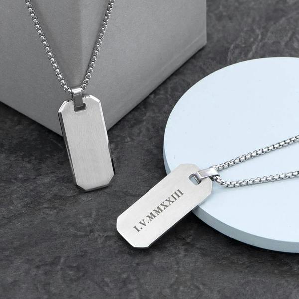Two personalised men's brushed steel dog tag necklaces on box chains, with one pendant showing a blank surface and the other engraved with Roman numerals.