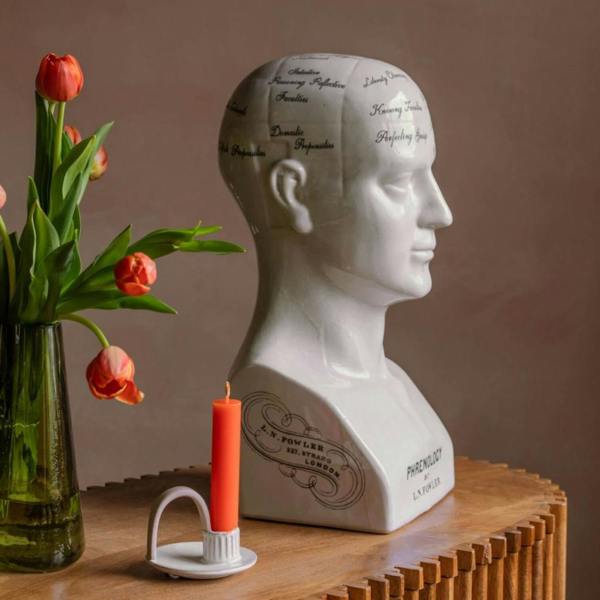 A large white porcelain phrenology head featuring black character-mapping text, sitting on a wooden surface next to a bright orange candle in a modern holder and a green glass vase filled with fresh red tulips.