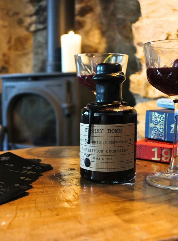 A dark glass bottle labeled 'Cherry Bomb' by Prohibition Cocktails sits on a wooden table in a cozy indoor setting with a candle, stone wall, and filled cocktail glasses nearby.
