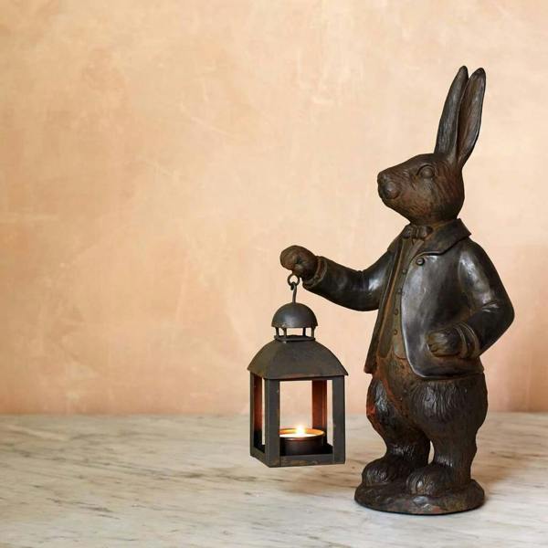 A dark bronze-coloured rabbit statue dressed in a suit holding a small black metal lantern with a lit tealight inside.