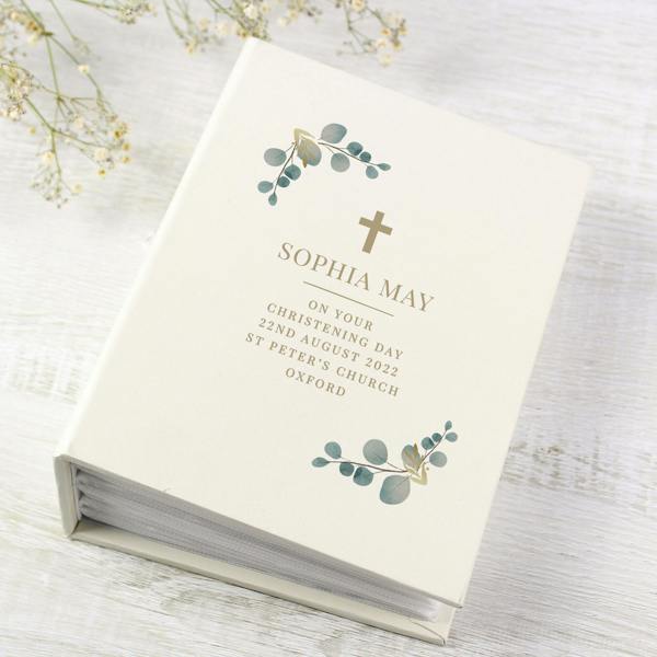 An ivory hardback 6x4 photo album by Abode Home & Gifts lying on a whitewashed wooden background next to white baby's breath flowers. The cover features a gold cross emblem, green eucalyptus foliage, and custom printed text that reads "SOPHIA MAY ON YOUR CHRISTENING DAY 22ND AUGUST 2022 ST PETER'S CHURCH OXFORD".