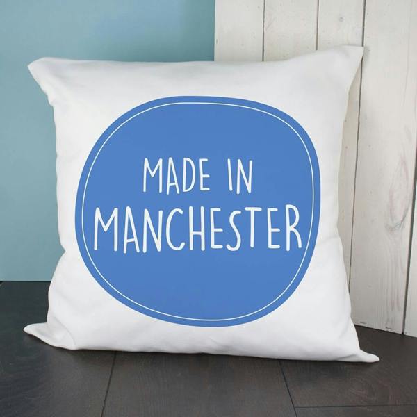 A square white poly-cotton cushion cover printed with a large blue oval graphic containing the white text MADE IN MANCHESTER.